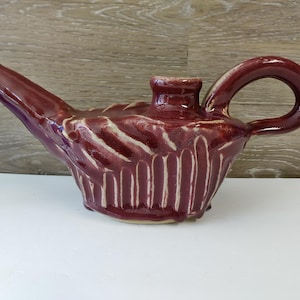May include: A maroon ceramic oil lamp with a long spout and a curved handle. The lamp has a textured surface with vertical and horizontal carved lines. The lamp is sitting on a white surface.