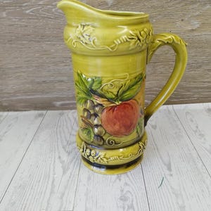 May include: A tall, olive-green ceramic pitcher with a curved handle and spout. The pitcher features a painted design of a red apple, grapes, and leaves. Decorative relief patterns of leaves and vines adorn the top and bottom.