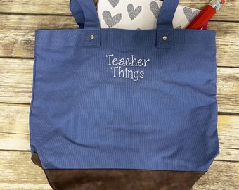 May include: Blue canvas tote bag with brown leather handles and trim. The bag has the text "Teacher Things" embroidered on the front in white.