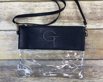 Puede incluir: Bolso bandolera negro con frontal transparente. El bolso presenta una sección superior negra con las letras "GT" en relieve y cremallera y herrajes de correa dorados. La correa ajustable es negra.