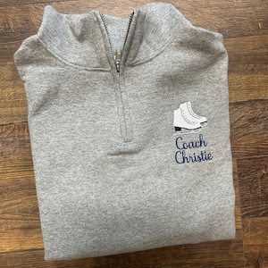 以下が含まれることがあります： グレーのクォータージップスウェットシャツに、白いスケートと「コーチクリスティー」の文字がネイビーブルーで刺繍されています。
