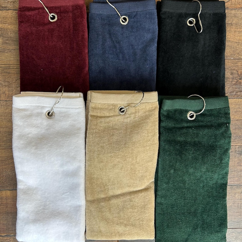 May include: A collection of golf towels in various colors, including burgundy, navy blue, black, white, tan, and forest green. Each towel is folded and has a metal hook for hanging. The towels are made of a soft, absorbent material.