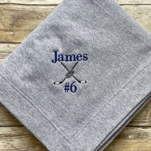 以下が含まれることがあります： グレーのフリースブランケットに、青い刺繍で「James #6」とホッケーのスティックとパックのデザインが施されています。