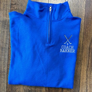 Puede incluir: Sudadera azul real con cremallera de un cuarto con un logotipo bordado en blanco que dice "COACH BAKNER" y presenta dos palos de hockey cruzados con un disco en el medio.