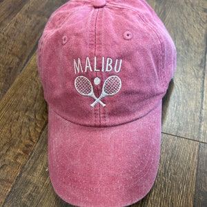 May include: A dusty rose baseball cap with the word "MALIBU" embroidered in white above a tennis ball and crossed rackets. The cap is made of a soft, worn fabric.