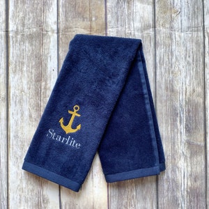 May include: A navy blue towel featuring a gold anchor design and the name "Starlite" embroidered on it. The towel is folded and displayed on a wooden surface. The towel has a dark blue border.