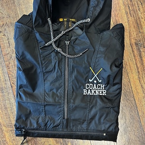 May include: Black hooded windbreaker jacket with a zipper closure and a white and black drawstring. The jacket has a logo embroidered on the front that says "Coach Bakner" with two crossed golf clubs.