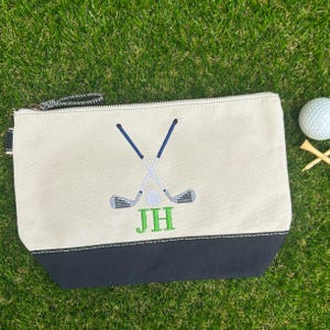 May include: A beige canvas zippered pouch with a black bottom panel. The front features an embroidered design of two crossed golf clubs in navy blue and grey, a small white golf ball, and the green embroidered initials JH centered below.