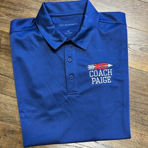 May include: A blue polo shirt with the text "PORT AUTHORITY" and "M" on the collar. The shirt has an embroidered logo with the text "XC COACH PAIGE" in red and white.