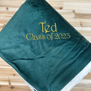 May include: A green fleece blanket with a white sherpa lining. The blanket has gold embroidery that reads "Ted Class of 2023".