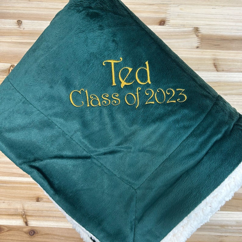 May include: A green fleece blanket with a white sherpa lining. The blanket has gold embroidery that reads "Ted Class of 2023".