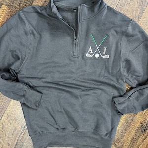 Puede incluir: Una sudadera gris oscuro con cremallera de un cuarto de vuelta con un diseño bordado en blanco de dos palos de golf cruzados y una bola de golf con las iniciales "AJ" sobre los palos.