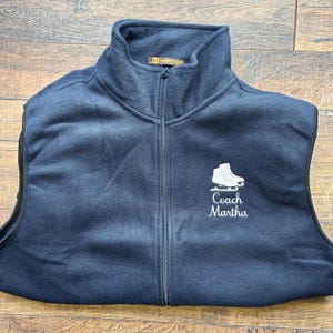 May include: Navy blue fleece vest with a full zipper and a high collar. The vest features a white embroidered ice skate and the text "Coach Martha". The vest has the "H Harrington" logo on the collar.