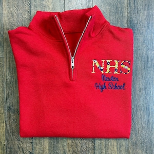 May include: A red quarter-zip sweatshirt. The letters "NHS" are embroidered in a floral pattern, with "Newton High School" below in navy blue. The sweatshirt has a silver zip and is neatly folded.