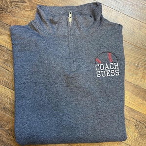 May include: A gray quarter-zip sweatshirt with a baseball graphic and the text "COACH GUESS" embroidered in white. The baseball design features red stitching. The sweatshirt has a high collar and a silver zipper.
