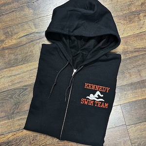 May include: Black zippered hoodie with a white swimmer graphic and the text "KENNEDY SWIM TEAM" in orange.