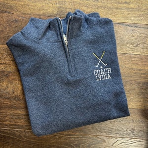 Puede incluir: Una sudadera azul oscuro con cremallera de un cuarto de vuelta con un logotipo bordado en blanco que dice "COACH LYDIA" y un par de palos de golf cruzados con una bola de golf.