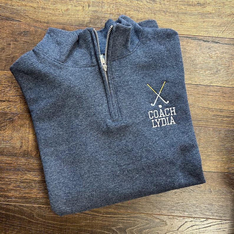 Puede incluir: Una sudadera azul oscuro con cremallera de un cuarto de vuelta con un logotipo bordado en blanco que dice "COACH LYDIA" y un par de palos de golf cruzados con una bola de golf.
