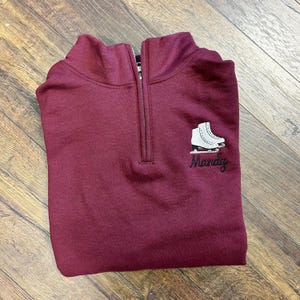 以下が含まれることがあります： ワインレッドのクォータージップスウェットシャツ。白いスケートのグラフィックと、黒で刺繍された「Mandy」という名前がデザインされています。