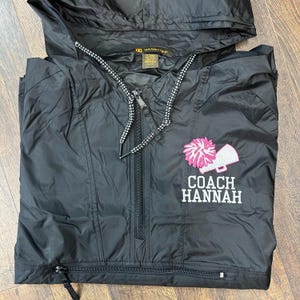 May include: Black hooded jacket with a half-zip closure and drawstring. Features a pink and white pom-pom and megaphone design with the text "COACH HANNAH" embroidered in white. The jacket has a small zipper pocket and a black zipper.