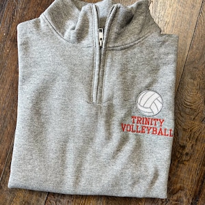 Peut inclure: Sweat-shirt gris à fermeture éclair sur le devant avec un ballon de volley brodé en rouge et le texte "Trinity Volleyball" sur le côté droit.