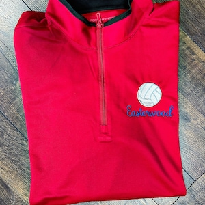 Op de afbeelding: Een rode sporttop met een kwartrits, zwarte kraag en een wit en blauw volleybal logo. Het woord "Easterwood" is eronder in blauwe letters geborduurd. De top is gemaakt van een glad, kreukvrij materiaal.