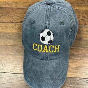 Könnte beinhalten: Eine dunkelblaue Baseballkappe mit einem weißen Fußball und dem Wort "COACH" in Gelb gestickt.