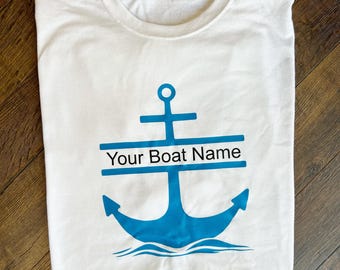 May include: White t-shirt with a blue anchor design. The anchor has a horizontal bar with the text "Your Boat Name" above the anchor's shaft. The design includes a wave at the bottom of the anchor.