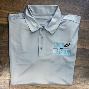 May include: A light gray polo shirt with a three-button placket and a collared neckline. The shirt has the text "COACH BRAYLYNN" embroidered in teal. The brand "PORT AUTHORITY" is printed on the inside collar.