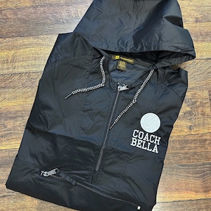 May include: Black hooded windbreaker jacket with a white embroidered logo that says "Coach Bella" and a white basketball graphic.