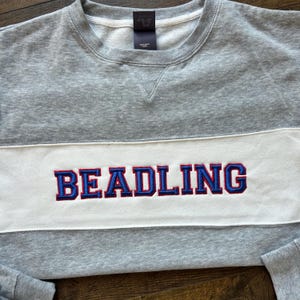 May include: A gray sweatshirt with a white horizontal stripe across the chest. The word "BEADLING" is embroidered in blue with red trim on the white stripe. The sweatshirt has a crew neck and long sleeves.