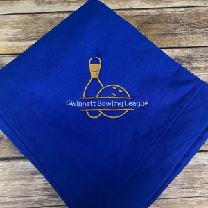 May include: A royal blue fleece blanket with a gold embroidered bowling pin and ball with the text "Gwinnett Bowling League" in a gold frame.