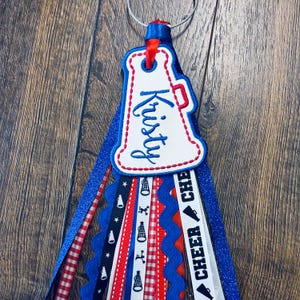 Op de afbeelding: Een cheerleading megafoon sleutelhanger met een witte geborduurde patch met de naam Kristy in blauw schrift, bevestigd aan een metalen ring met meerdere hangende linten in rood, wit en blauw, inclusief glitter, vichy en megafoon prints.