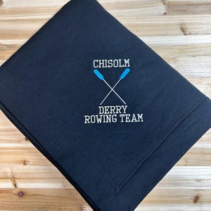 May include: Black fleece throw blanket with white embroidered text "CHISOLM DERRY ROWING TEAM" and two blue oars crossed in the centre.