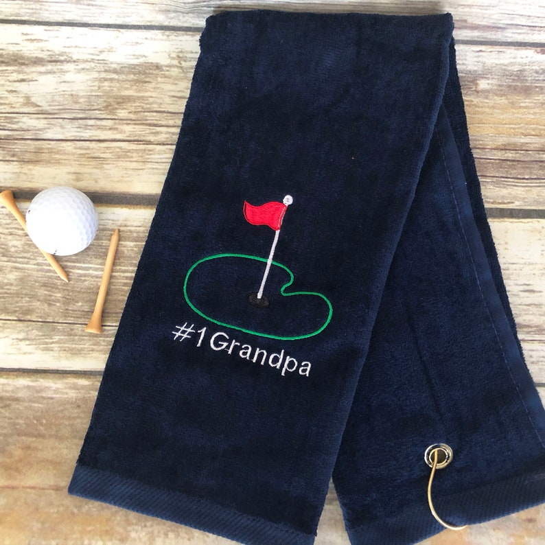 May include: Navy blue golf towel with embroidered design. Features a red flag on a green putting green, with the text "#1 Grandpa" in white. Includes a golf ball and tees.