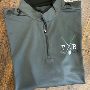 May include: A gray golf shirt with a black zipper and a green and white golf club design with the letters "TXB" on the front.