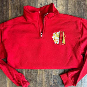 Könnte beinhalten: Ein rotes, verkürztes Sweatshirt mit hohem Kragen und einem Viertel-Reißverschluss. Das Sweatshirt hat ein gesticktes Design aus einem gelb-weißen Pompon und dem Wort "Mustangs" in goldfarbenen Buchstaben.