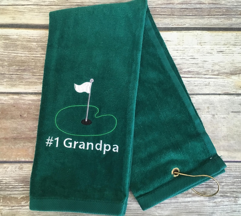 May include: A folded, dark green golf towel with the embroidered image of a golf hole and flag. The text "#1 Grandpa" is embroidered below the image in white. A gold-colored metal loop is attached to the corner of the towel.