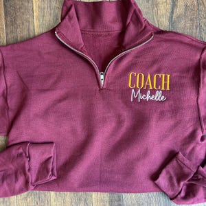 Peut inclure: Un sweat-shirt zippé quart de tour de couleur bordeaux avec un col montant et des manches longues. Le texte "COACH Michelle" est brodé en jaune et blanc sur le devant. La fermeture éclair est argentée.