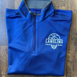 Op de afbeelding: Een koningsblauwe sporttrui met een kwartrits en een grijze kraag. De trui heeft de witte tekst "LANSLING BASKETBALL", een basketbalafbeelding en een gestileerd "W"-logo. De rits loopt over de borst.