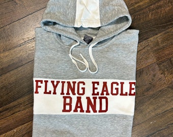 Puede incluir: Sudadera gris con capucha, con una franja blanca en la espalda y una banda blanca en la parte delantera. La banda tiene las palabras "FLYING EAGLE BAND" en letras rojas.