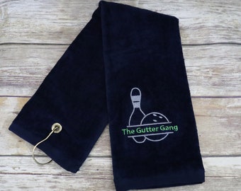 May include: A navy blue towel with a white embroidered bowling pin and ball design. The text "The Gutter Gang" is embroidered in green below the bowling pin and ball.