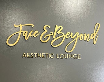 May include: A gold sign on a gray wall reads "Face & Beyond Aesthetic Lounge".