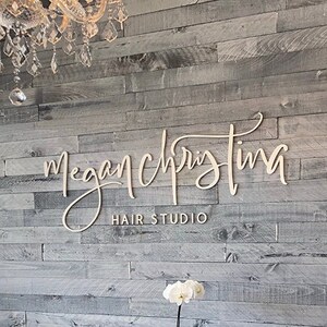 K&ouml;nnte beinhalten: Ein Wartezimmer in einem Friseursalon mit zwei braunen Ledersesseln, einem kleinen Tisch mit einer Zeitschrift und einer wei&szlig;en Orchideenpflanze. Der Name des Salons, "Megan Christina Hair Studio", ist an einer grauen Holzwand angebracht.