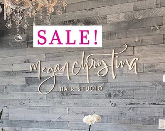 May include: A gray wood-paneled wall with a white sign that reads "Megan Christina Hair Studio" and the word "SALE!" in pink letters. Two brown leather chairs are in front of the wall, and a white orchid plant is on a table between the chairs.