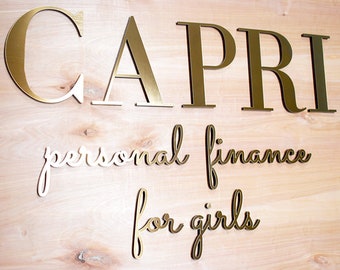 May include: Gold-colored letters spelling "CAPRI" above the words "personal finance for girls" on a light wood background. The letters are in a bold, sans-serif font, while the other text is in a cursive script.