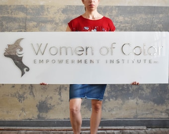 May include: A white sign with the text "Women of Color Empowerment Institute, Inc." in silver lettering. A silver stylized female face is on the left side of the sign.
