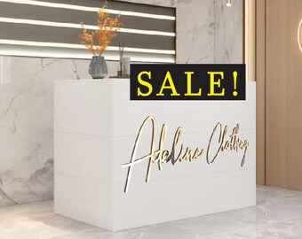 May include: A white counter with a gold inscription that reads "Atelina Clothing" and a black sign above it that reads "SALE!"