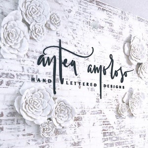 K&ouml;nnte beinhalten: Eine wei&szlig;e Backsteinwand mit einem schwarzen Schild, auf dem "Antea Amoroso Hand Lettered Designs" steht. Die Wand ist mit wei&szlig;en Papierblumen verziert.