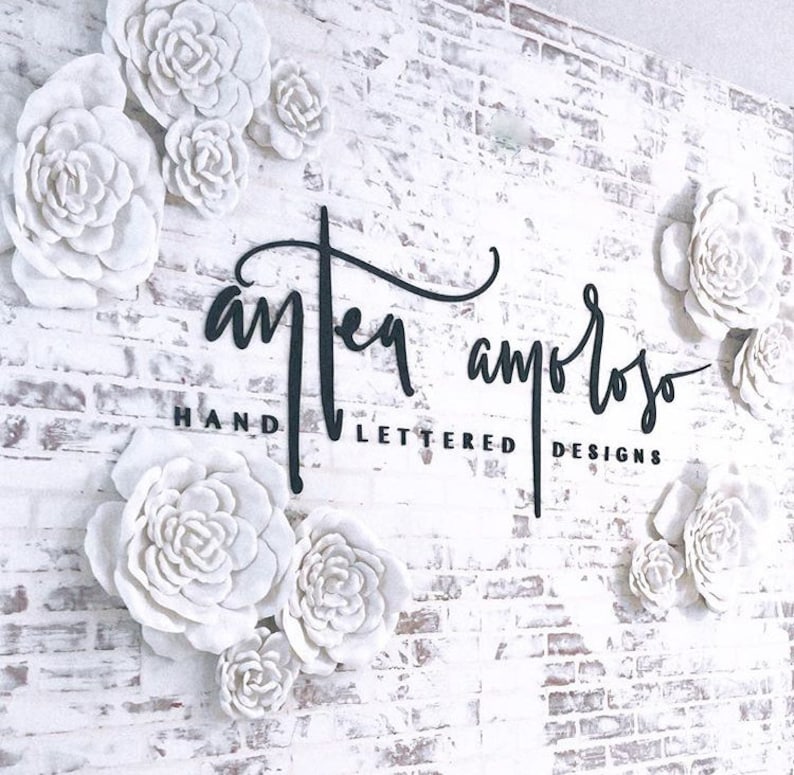 K&ouml;nnte beinhalten: Eine wei&szlig;e Backsteinwand mit einem schwarzen Schild, auf dem "Antea Amoroso Hand Lettered Designs" steht. Die Wand ist mit wei&szlig;en Papierblumen verziert.
